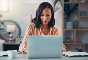 A woman with shoulder-length dark hair sits at a desk, smiling as she works on a laptop. An open notebook, coffee cup, and blurred background with a bicycle and shelves suggest she's enjoying her job.