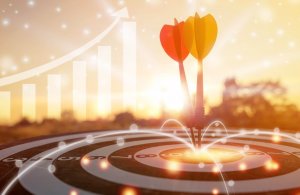 Two darts, one red and one yellow, hit the bullseye of a dartboard. In the background, a rising bar graph and upward arrow symbolize employee growth and success, set against a warm, glowing sunset.