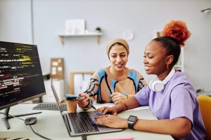 Two women sit at a desk, one wearing headphones, both smiling while looking at a laptop. A monitor displays colorful code as they collaborate in a modern office—perhaps developing solutions for governments.