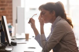 A woman sits at her desk in front of a computer, holding her glasses in one hand and rubbing her eyes with the other, appearing tired or stressed as she works on government documents.