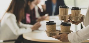 A person holds two trays with takeaway coffee cups in the foreground, while a group gathers at a round table in the blurred background, reflecting a collaborative office culture.