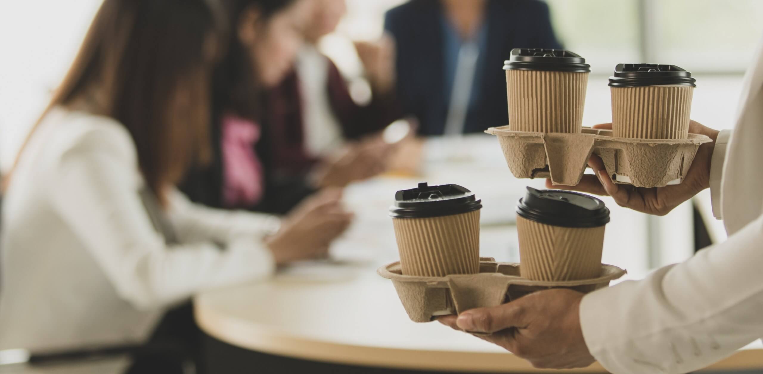 A person holds two trays with takeaway coffee cups in the foreground, reflecting a culture of collaboration as a group of people have a meeting around a table in the blurred background. A person holds two trays with takeaway coffee cups in the foreground, reflecting a culture of collaboration as a group of people have a meeting around a table in the blurred background.