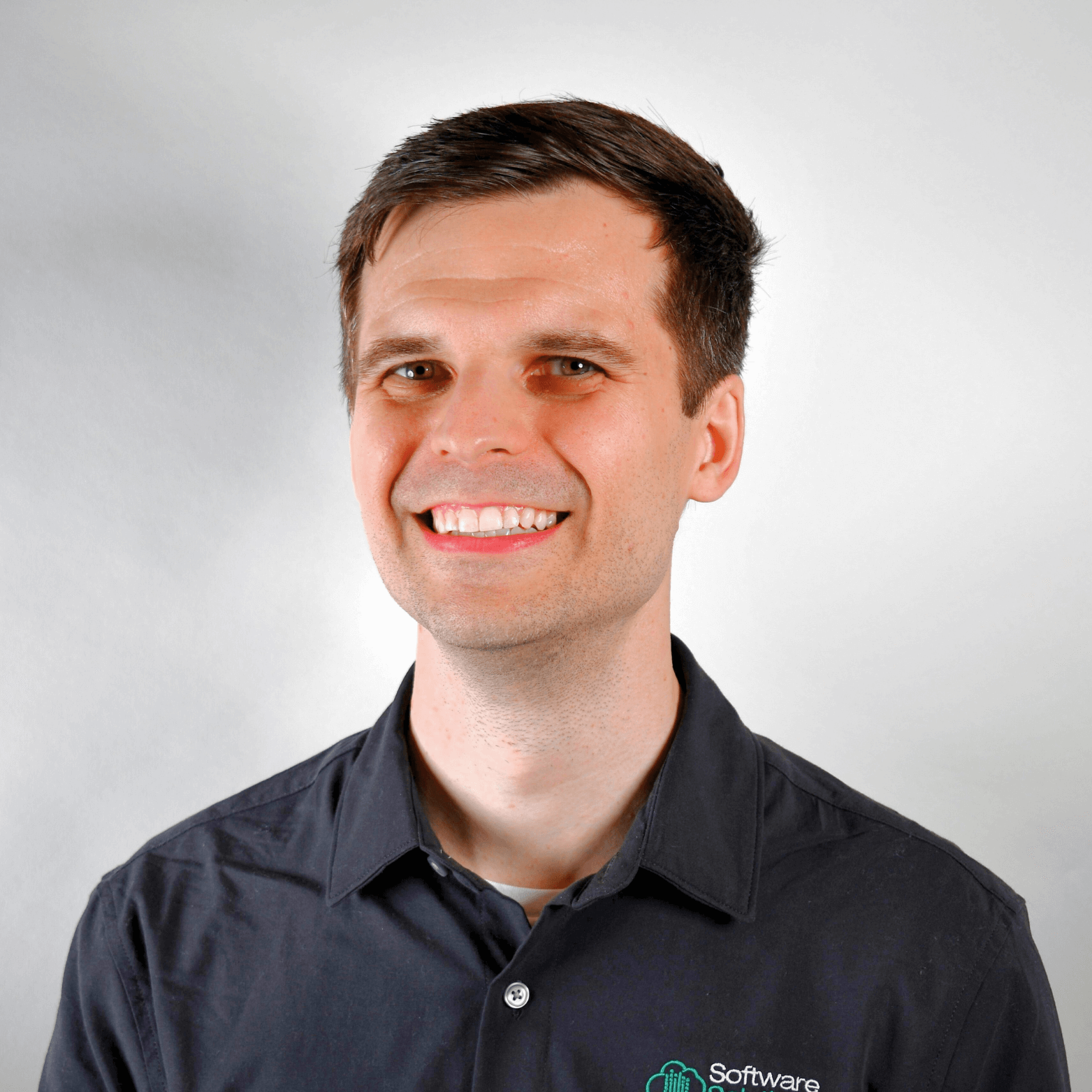A man with short brown hair smiles at the camera, wearing a black collared shirt with a small logo on the left side. The background is plain and light-colored. A man with short brown hair smiles at the camera, wearing a black collared shirt with a small logo on the left side. The background is plain and light-colored.