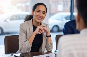 A woman in a blazer sits at a table, smiling and attentively listening to someone across from her during a government meeting or interview. Papers and a smartphone are visible on the table.