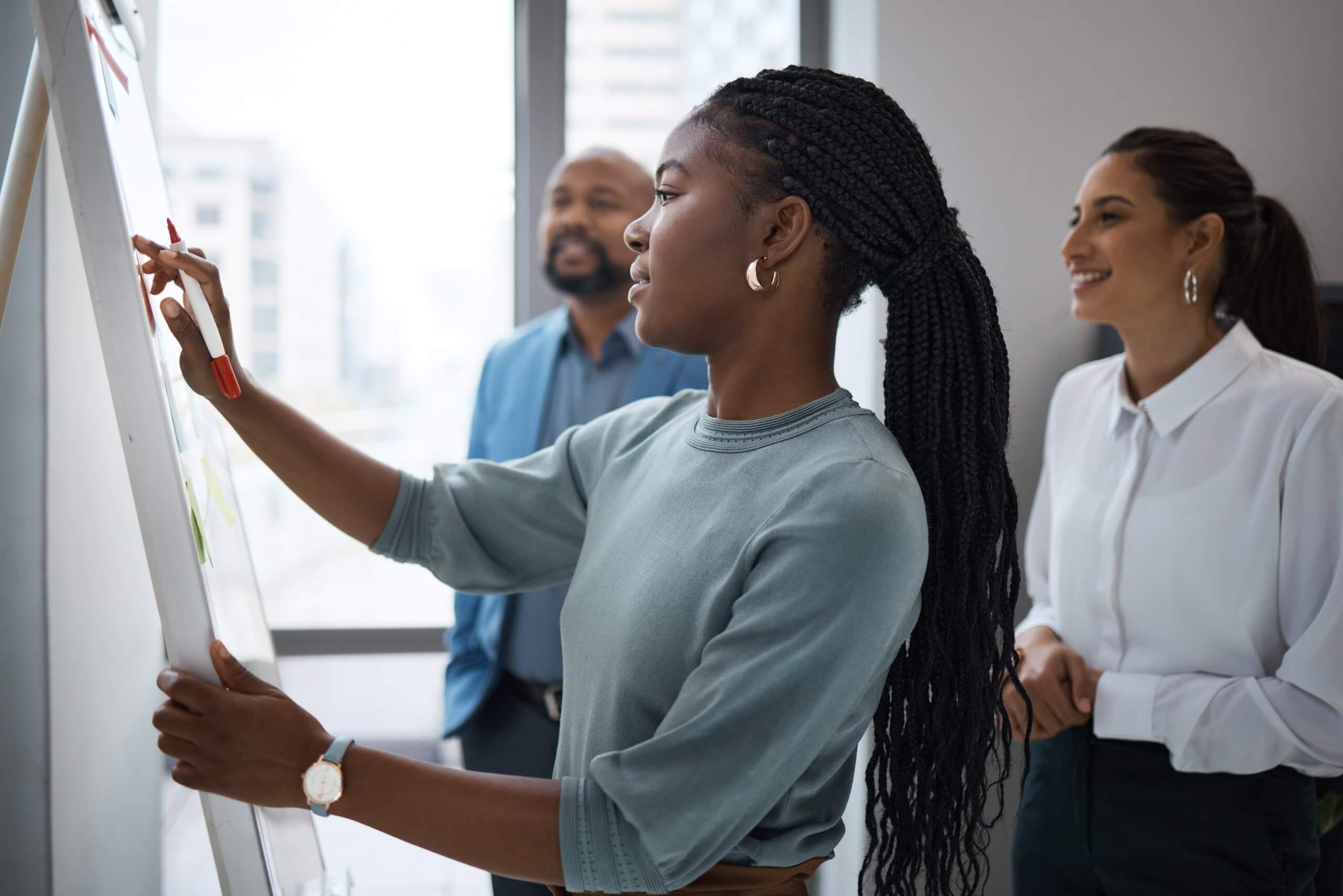 A woman writes on a flipchart while two colleagues, one man and one woman, stand beside her smiling in a bright government office setting. A woman writes on a flipchart while two colleagues, one man and one woman, stand beside her smiling in a bright government office setting.