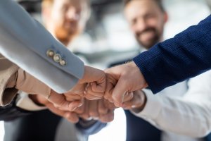 A group of people in business attire stand in a circle, smiling, as they bring their fists together for a group fist bump, symbolizing teamwork, unity, and strong leadership.
