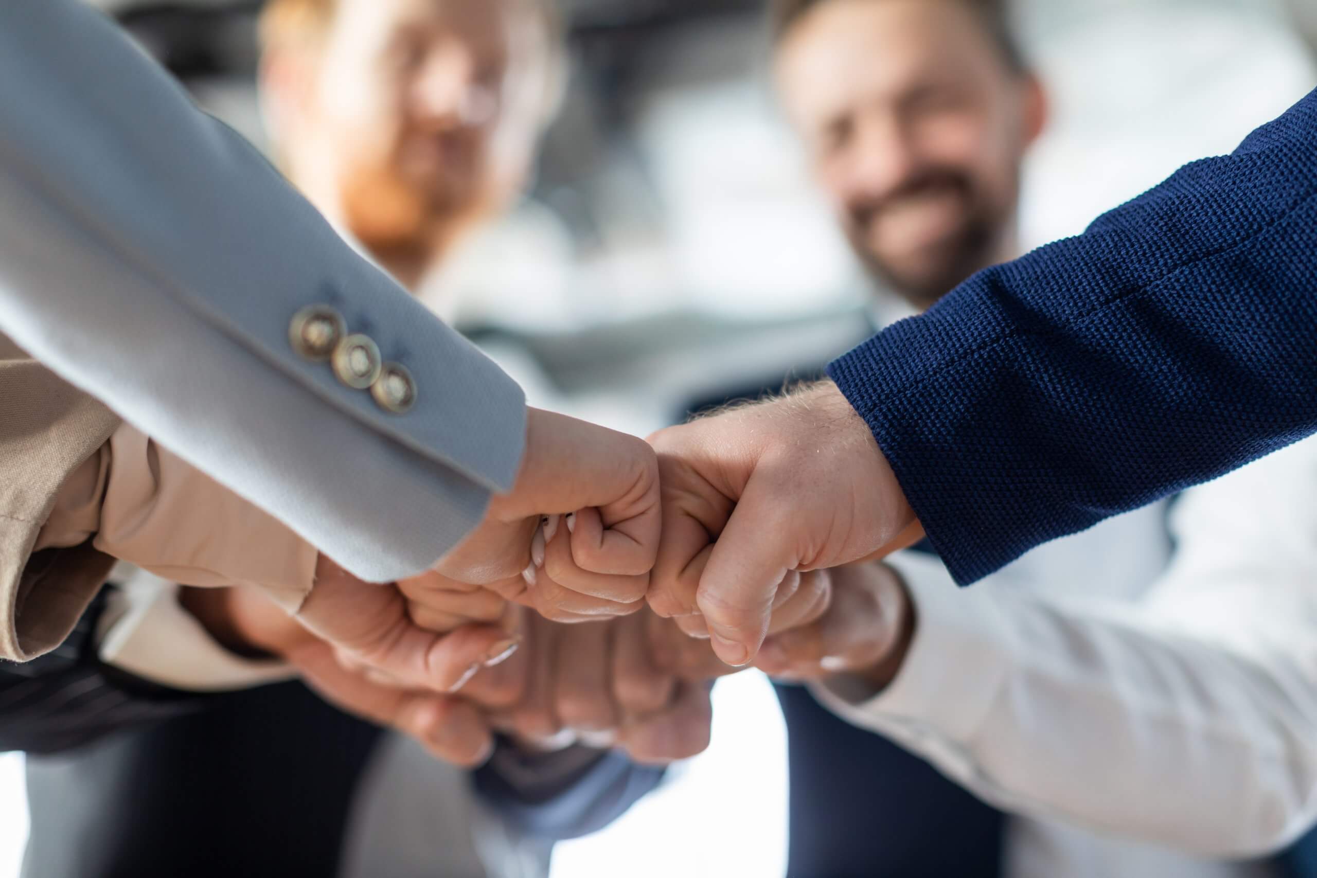 A group of people in business attire stand in a circle, smiling, as they bring their fists together for a group fist bump, symbolizing teamwork, unity, and strong leadership. A group of people in business attire stand in a circle, smiling, as they bring their fists together for a group fist bump, symbolizing teamwork, unity, and strong leadership.