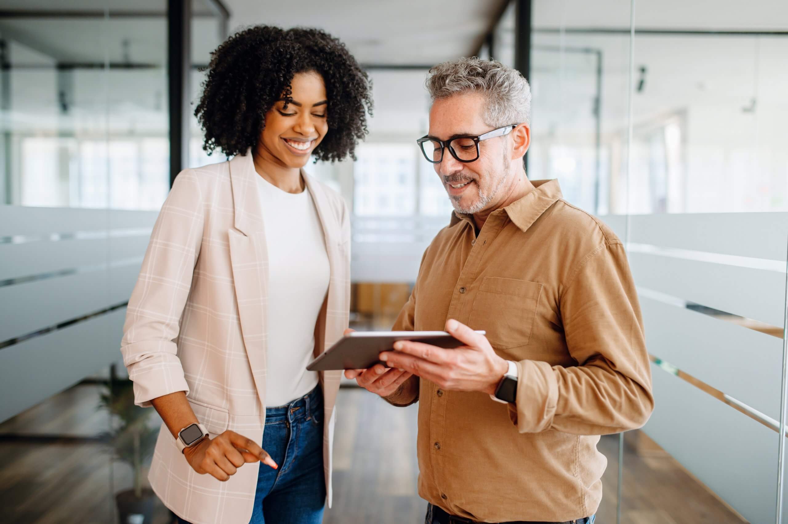 Two colleagues stand in a modern office hallway, smiling and looking at a tablet together. Their engaged discussion reflects teamwork and leadership, as they collaborate on new ideas for their project.