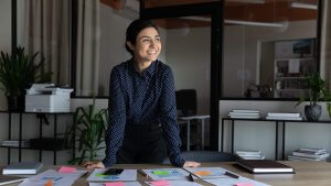 A woman in a dark polka dot blouse stands smiling with satisfaction at a desk covered with documents, charts, and colorful sticky notes in a modern office with plants and shelves in the background.