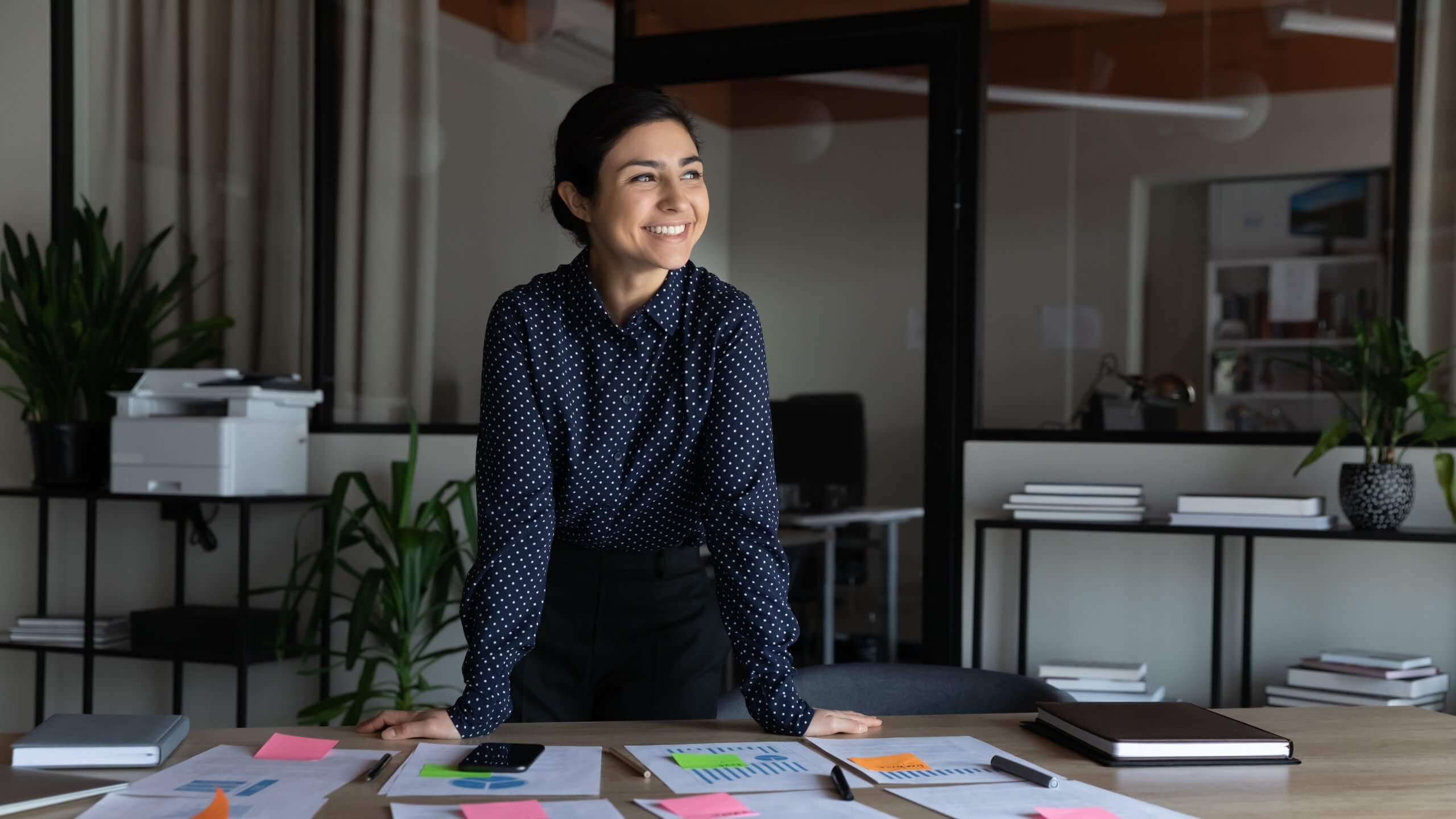 A woman in business attire stands smiling at her desk, confidently managing job-related papers, charts, and sticky notes in a modern office with large windows, plants, and shelves in the background. A woman in business attire stands smiling at her desk, confidently managing job-related papers, charts, and sticky notes in a modern office with large windows, plants, and shelves in the background.