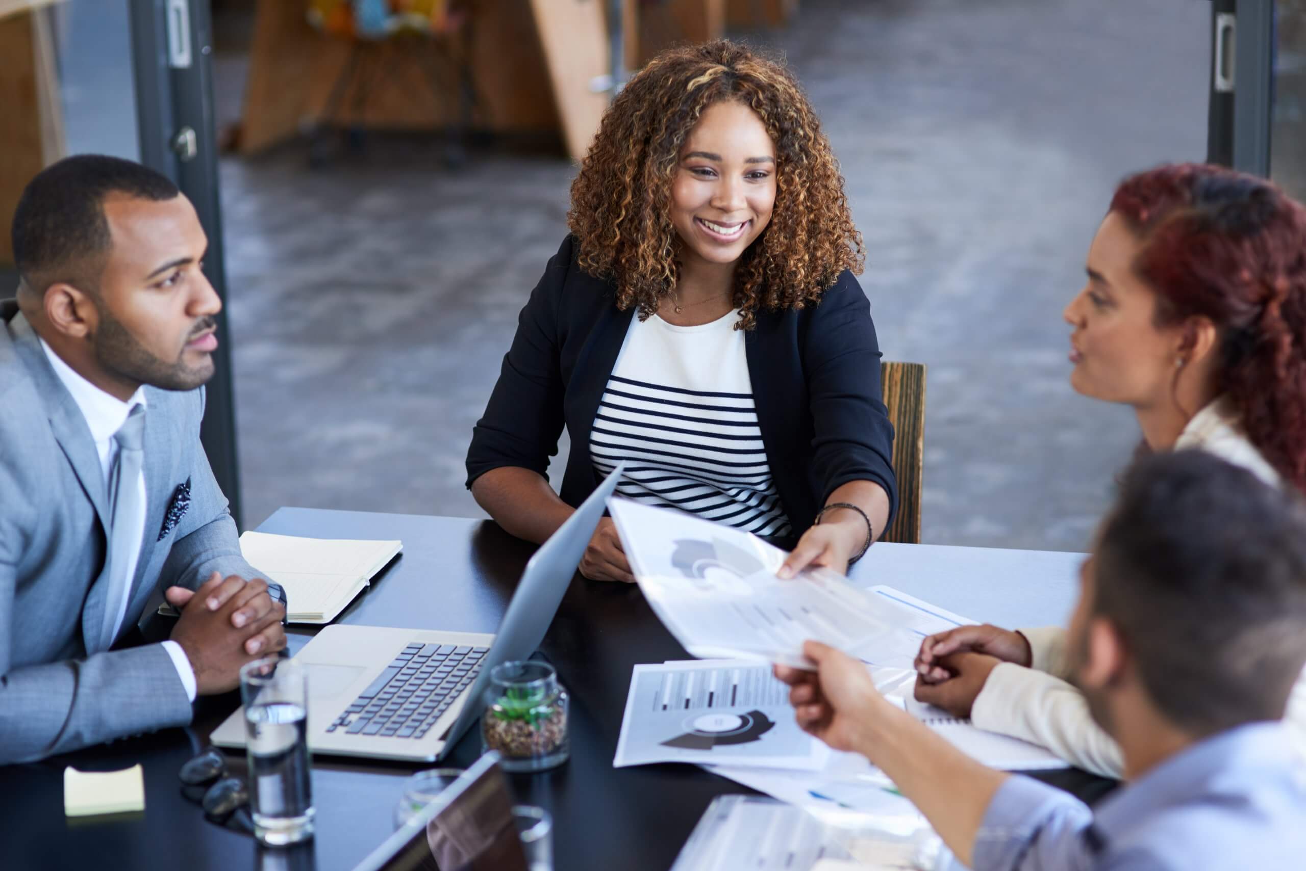 Four employees sit around a table in an office, discussing documents and charts. One woman is smiling as she hands a paper to another person. A laptop, glasses of water, and papers are on the table. Four employees sit around a table in an office, discussing documents and charts. One woman is smiling as she hands a paper to another person. A laptop, glasses of water, and papers are on the table.