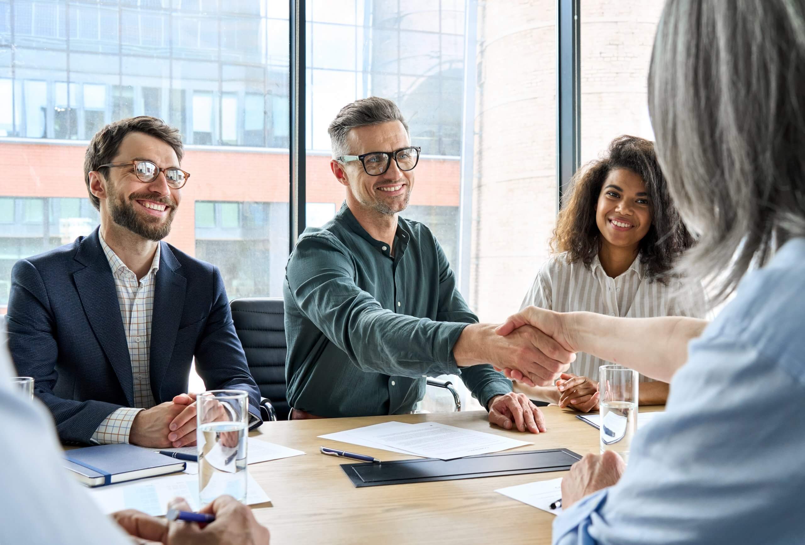 Four people in business attire sit around a table in a modern office, with two in the center smiling and shaking hands, suggesting a successful government meeting or agreement. Papers, notebooks, and glasses of water are on the table. Four people in business attire sit around a table in a modern office, with two in the center smiling and shaking hands, suggesting a successful government meeting or agreement. Papers, notebooks, and glasses of water are on the table.