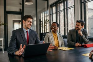 Three professionals in business attire sit around a table in a modern office, engaged in discussion. The group’s collaborative spirit reflects a positive company culture. A laptop and folders are on the table.