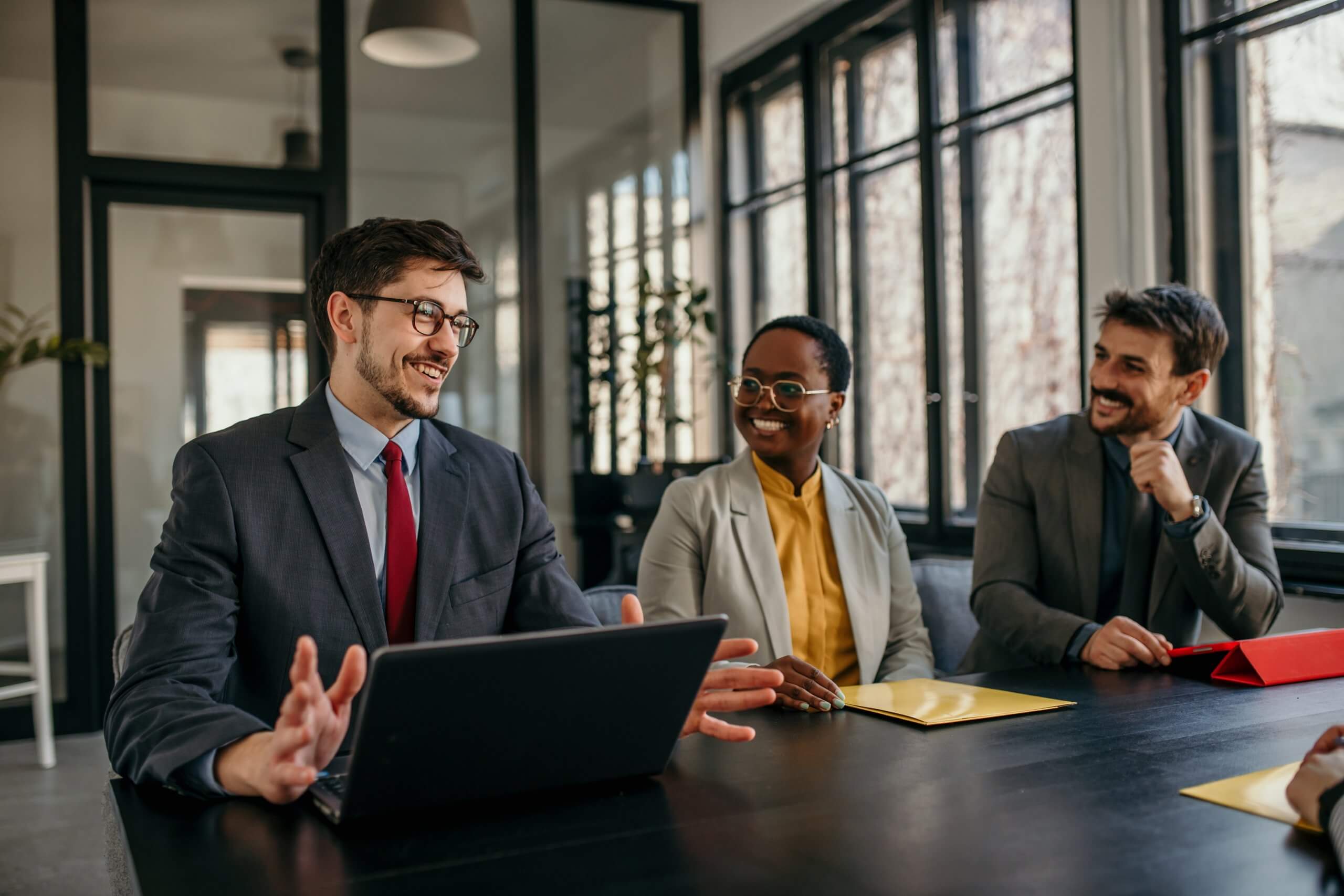 Three professionals sit at a conference table in a modern office, engaged in conversation. Demonstrating leadership, one man speaks while gesturing with a laptop in front of him, as the others listen and smile. Three professionals sit at a conference table in a modern office, engaged in conversation. Demonstrating leadership, one man speaks while gesturing with a laptop in front of him, as the others listen and smile.
