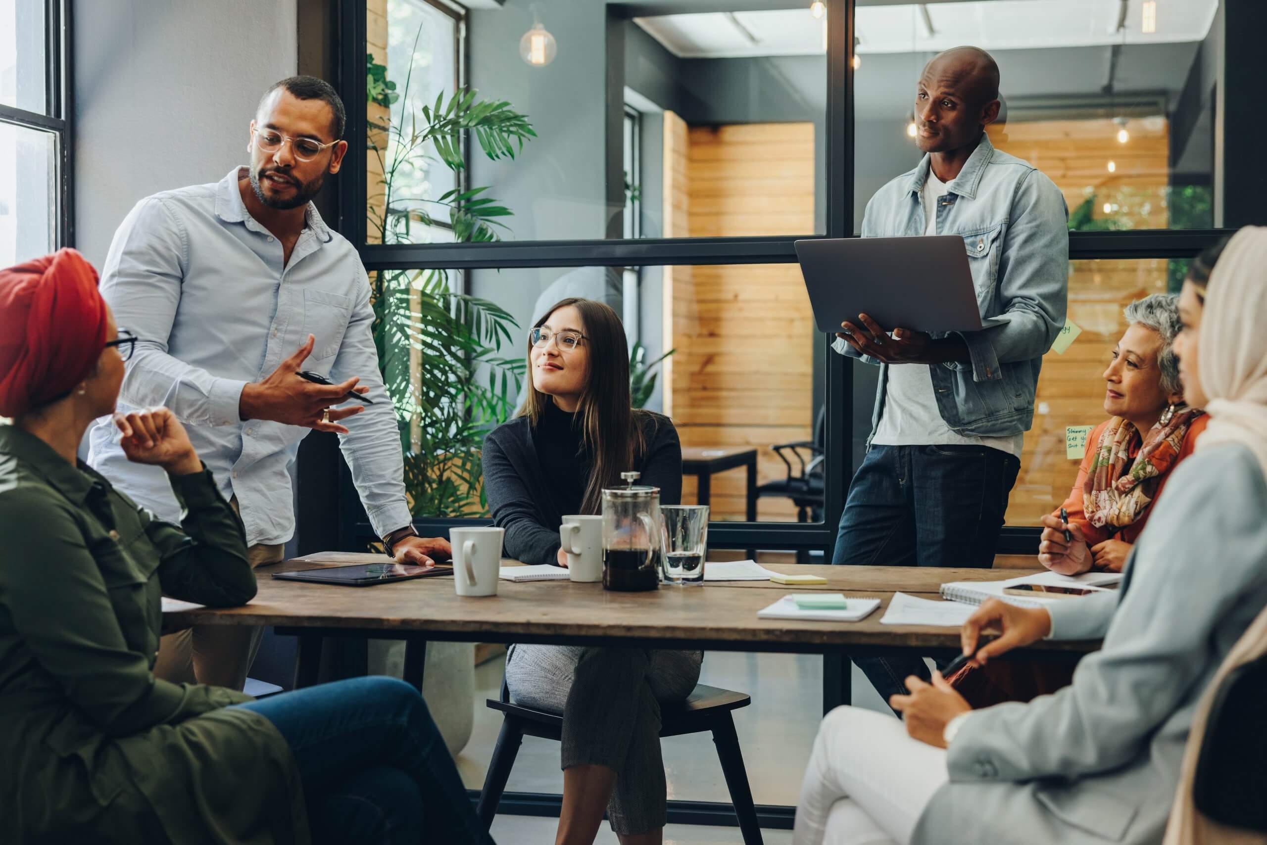 A group of six diverse people in casual attire are having a discussion around a table in a modern office with large windows, demonstrating leadership as one person stands with a laptop while others are seated and listening attentively.