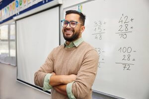 A smiling man with glasses and a beard stands with arms crossed, radiating satisfaction, in front of a whiteboard displaying math problems in a classroom.