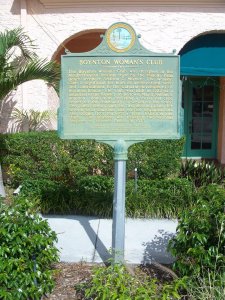 A green and gold historical marker for the Boynton Woman’s Club stands on a sidewalk with bushes and a pink building in the background, partially shaded by palm leaves.