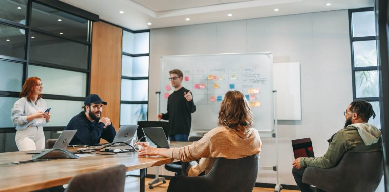 Five people are in a modern meeting room. One person stands at a whiteboard covered with sticky notes, presenting ideas for a public sector project, while the others sit at a table with laptops, listening and interacting.
