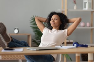 A woman sits at her desk with her legs up and hands behind her head, eyes closed, appearing relaxed after a long day on the job. Papers, a laptop, and office supplies cover the desk, with shelves and plants in the background.