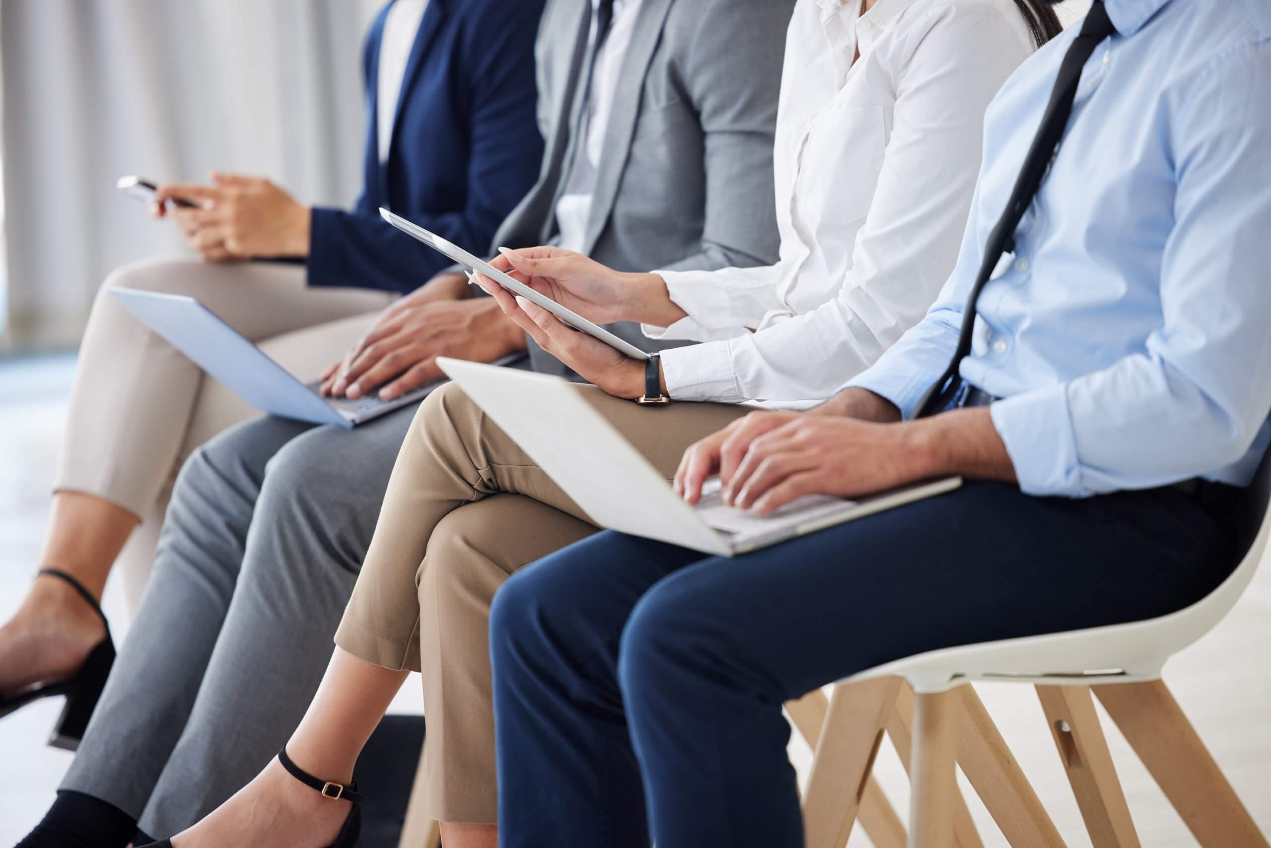 Four people in business attire sit in a row, using laptops, tablets, and smartphones—highlighting a professional, tech-savvy environment where AI seamlessly integrates into daily workflows. Only their lower bodies are visible. Four people in business attire sit in a row, using laptops, tablets, and smartphones—highlighting a professional, tech-savvy environment where AI seamlessly integrates into daily workflows. Only their lower bodies are visible.