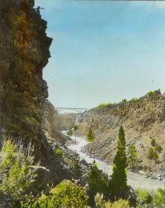 A scenic gorge with steep rocky cliffs, sparse vegetation, and a winding river below. In the background, a metal bridge spans the gap between the cliffs under a clear sky.
