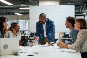 A group of five colleagues sit around a table in an office, engaged in a discussion. Demonstrating leadership, a man in a blue blazer stands, speaking enthusiastically as others listen and smile. A whiteboard with diagrams is in the background.
