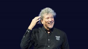 A smiling man with curly gray hair wearing a black shirt that says "GOOD COMES FIRST" gestures with his right hand against a plain dark background.