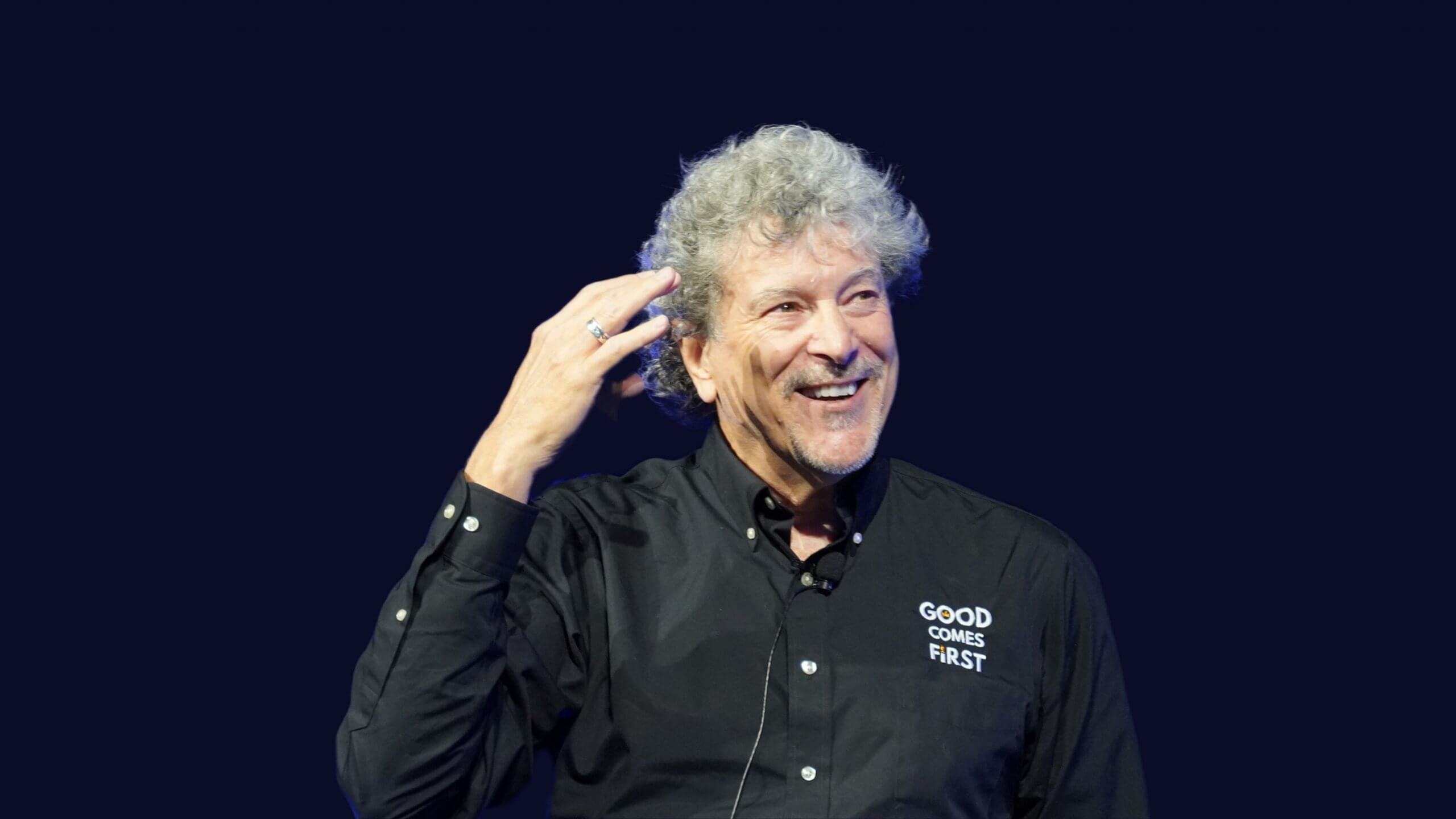 A smiling man with curly gray hair wearing a black shirt that says "GOOD COMES FIRST" gestures with his right hand against a plain dark background.