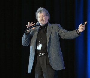 A man with gray curly hair and a beard, wearing a gray blazer and black shirt, speaks into a microphone on stage, gesturing with one hand while holding a device in the other. A lanyard hangs from his neck.