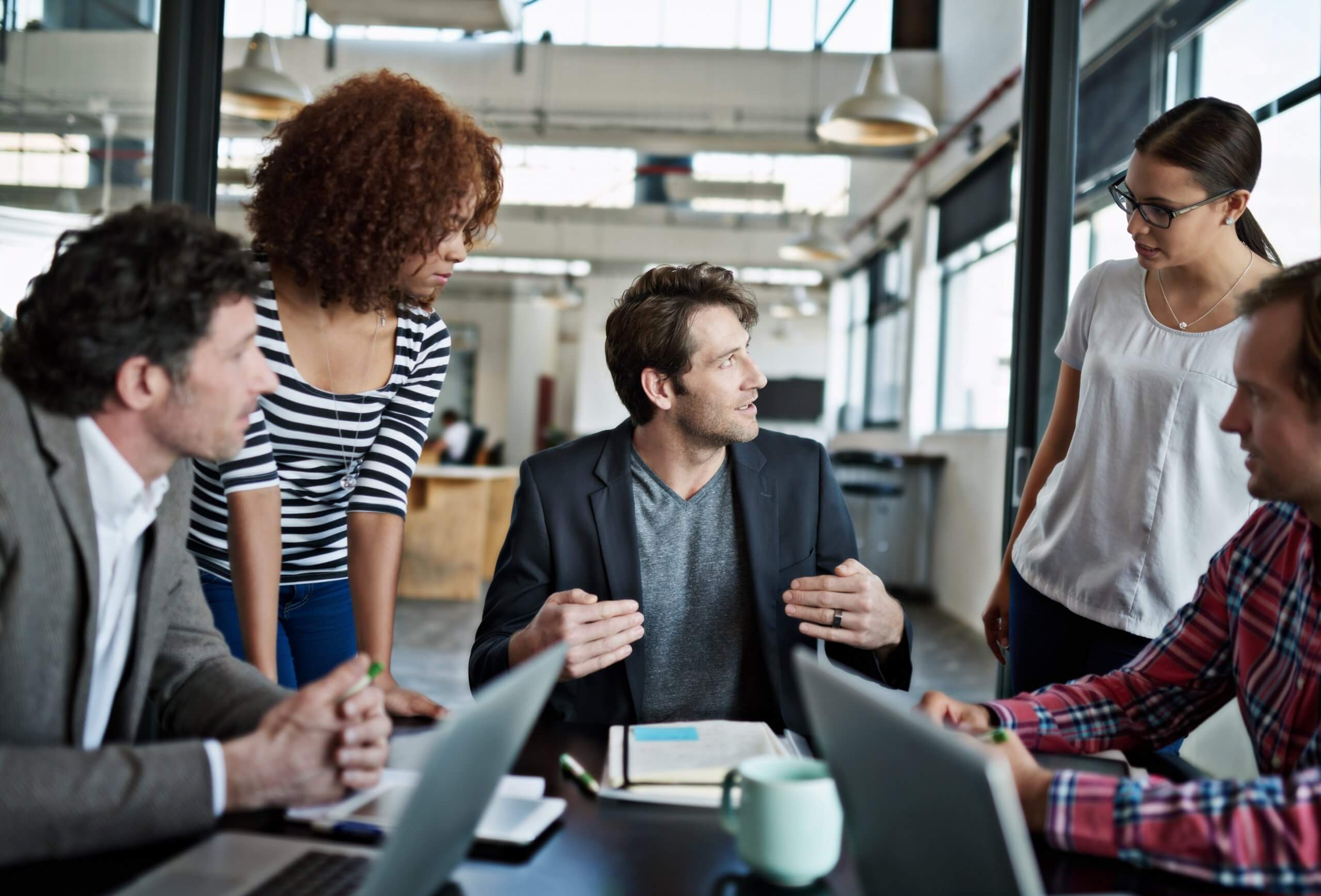 Four people stand and sit around a table in a modern office, having a discussion about public sector initiatives. Laptops, notebooks, and a coffee mug are on the table, while large windows fill the space with natural light.