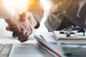 Two people shaking hands over a desk with documents, a pen, and a laptop, suggesting satisfaction and agreement in a professional setting with sunlight in the background.