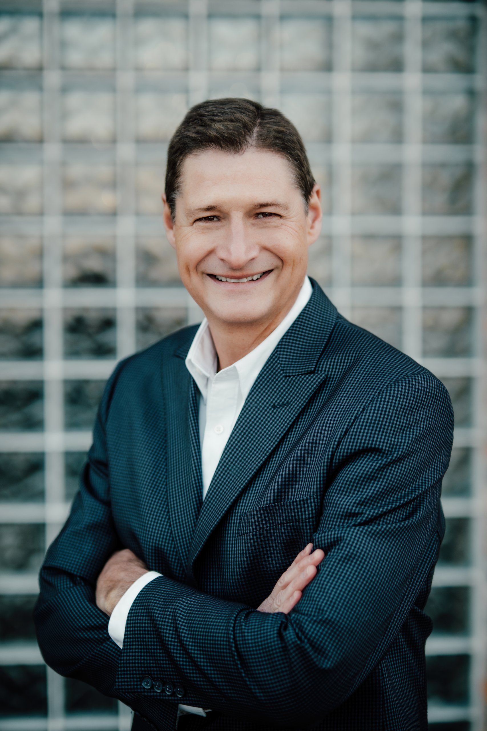 A man in a dark suit and white shirt stands with arms crossed, smiling confidently like today’s leaders, in front of a wall made of glass blocks. A man in a dark suit and white shirt stands with arms crossed, smiling confidently like today’s leaders, in front of a wall made of glass blocks.