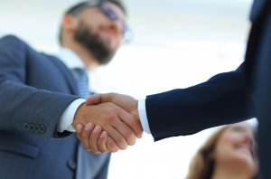 Two people in business suits shaking hands, viewed from a low angle. One person has a beard and glasses, and a woman is smiling in the blurred background, suggesting satisfaction with a new professional agreement or partnership.