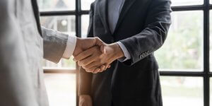 Two people in business suits shake hands in an office setting, symbolizing agreement or partnership and mutual satisfaction. A large window in the background lets in natural light.