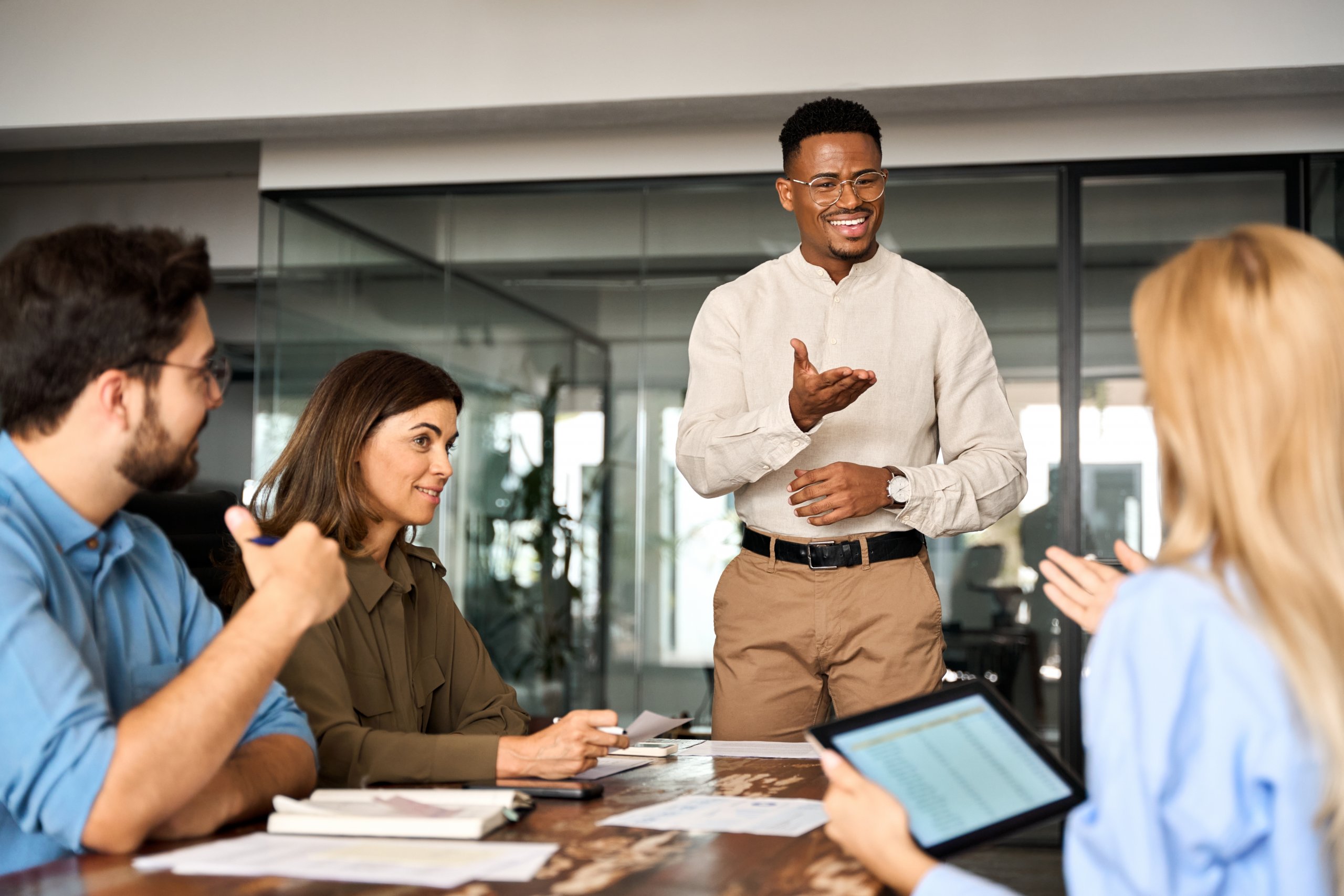 An employee stands and gestures while speaking to three colleagues seated at a conference table, engaged in discussion in a modern office setting with glass walls. An employee stands and gestures while speaking to three colleagues seated at a conference table, engaged in discussion in a modern office setting with glass walls.