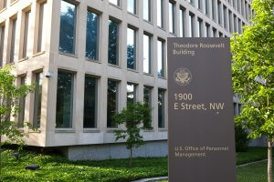 A sign outside a modern office building reads "Theodore Roosevelt Building, 1900 E Street, NW, U.S. Office of Personnel Management," hinting at a hub for career opportunities, with trees and windows visible in the background.