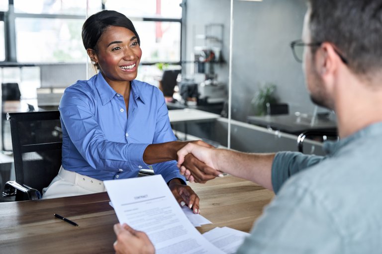 A smiling woman in a blue shirt shakes hands with a man across a desk, holding a contract. The modern office setting suggests a successful government job interview or business agreement.