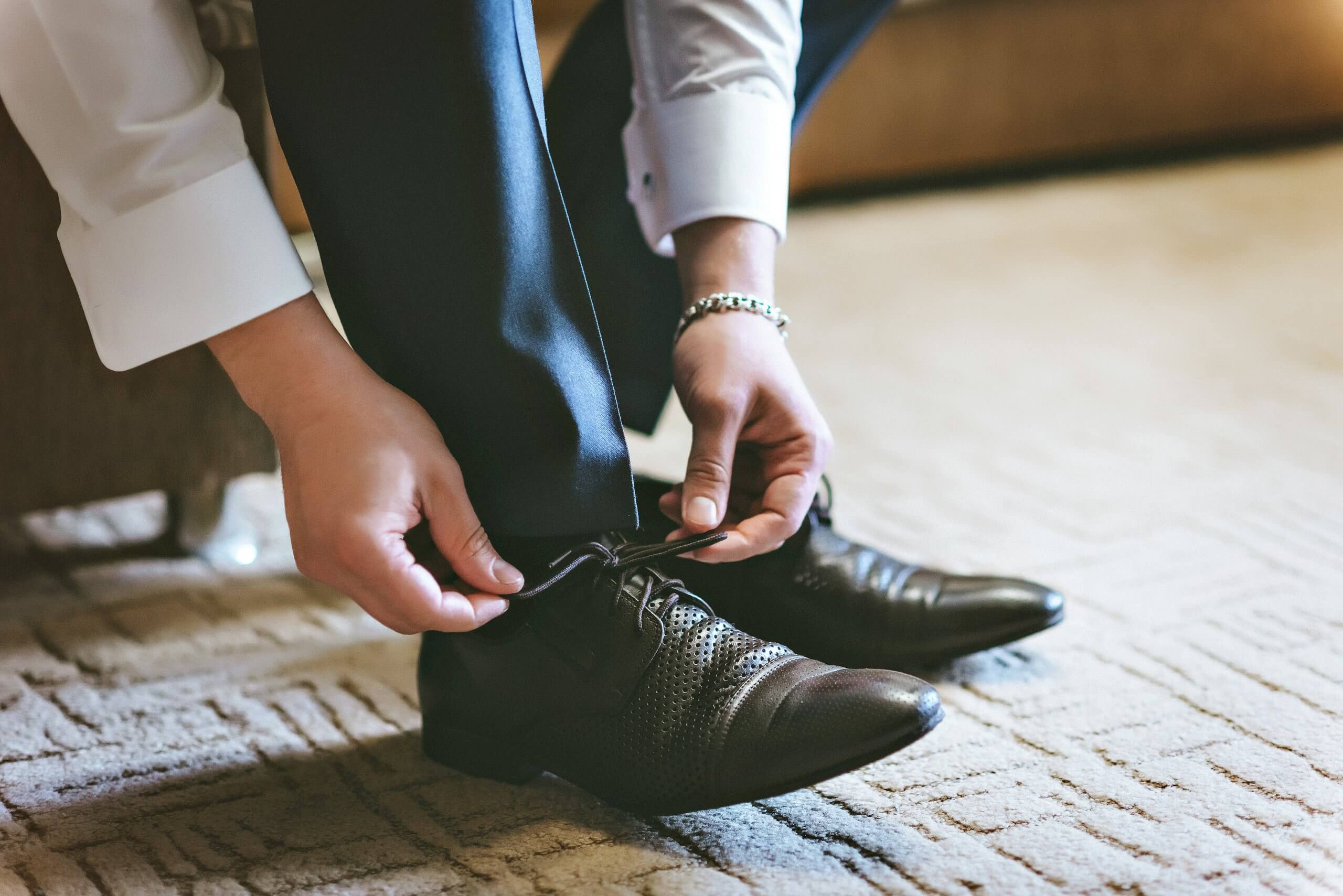 A leader in formal attire is sitting and tying the laces of black dress shoes on a patterned carpet, with only their hands and lower legs visible.