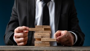 An employee in a suit stacks wooden blocks labeled with words like leadership, persistence, collaboration, strategy, adaptability, risk management, action, and planning on a table, representing building business skills.
