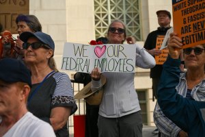 A group of people at a federal protest. One person in the center holds a sign that reads “We ♥ Our Federal Employees.” Others stand nearby with signs, all appearing focused and attentive.