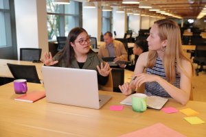 Two employees sit at a table with a laptop, notebooks, and mugs, deep in discussion. Two others work in the background of the bright, modern office space.
