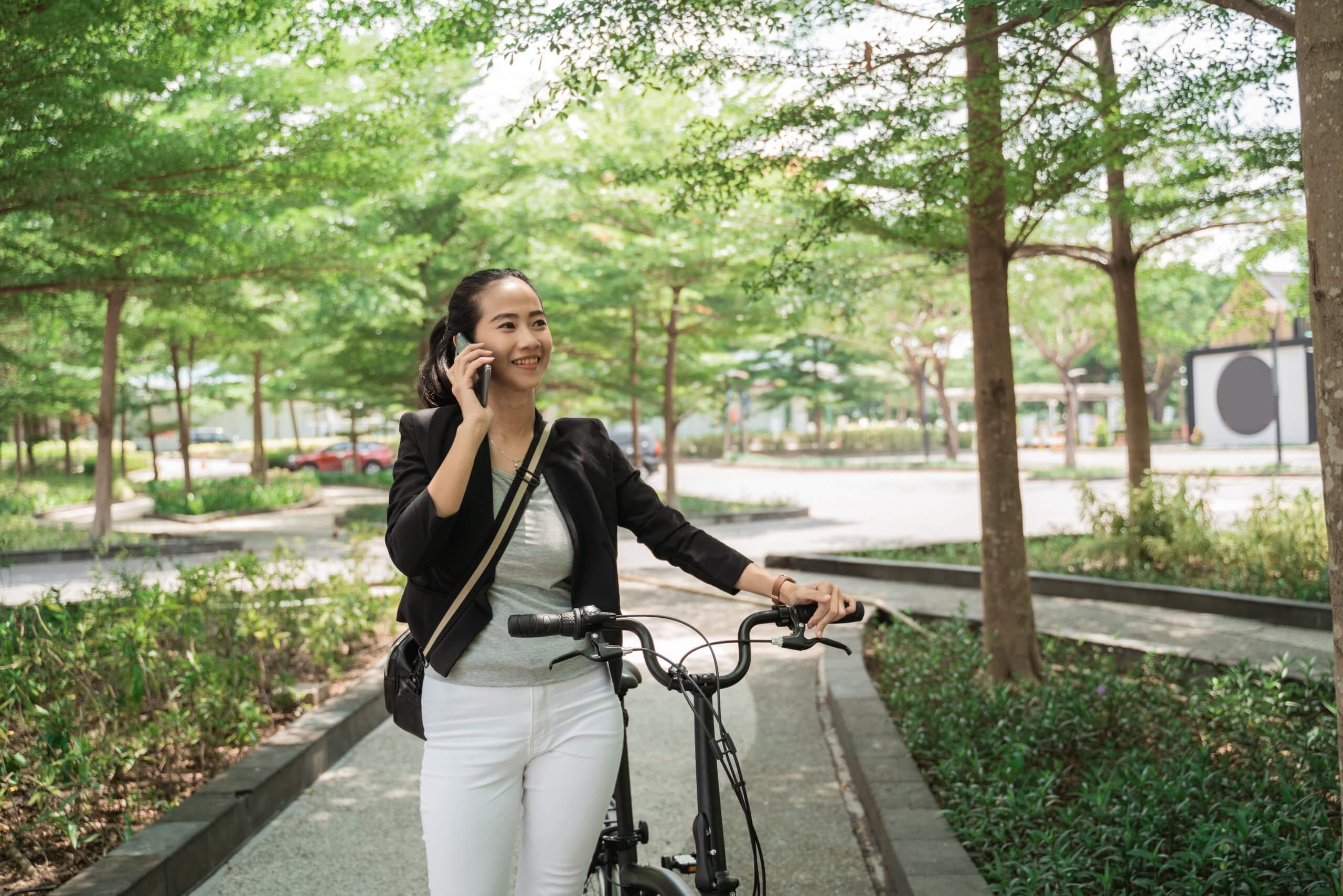 A woman, embodying the confidence of modern leaders, smiles while talking on her phone and walking her bicycle along a tree-lined path in a park on a sunny day. She wears a black blazer, white pants, and carries a crossbody bag.