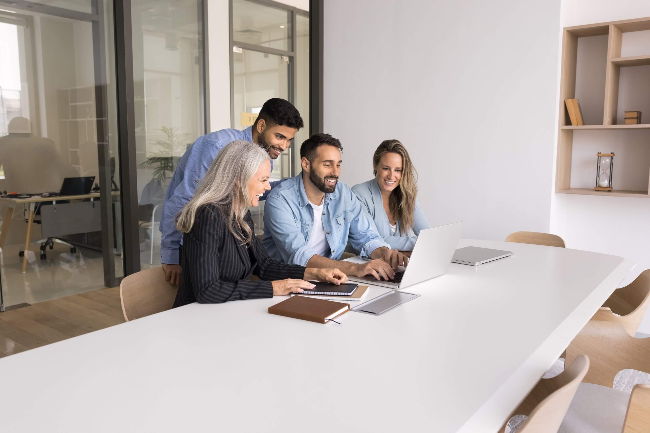 Multiethnic,Business,Team,Of,Positive,Different,Aged,Colleagues,Watching,Internet Four people in business attire sit around a table in a modern office, with two in the center smiling and shaking hands, suggesting a successful government meeting or agreement. Papers, notebooks, and glasses of water are on the table.