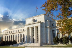 Large, white Federal-style stone building with tall columns and an American flag on top, bathed in sunlight with dramatic clouds and a tree with autumn leaves in the foreground.