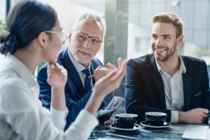Three businesspeople sit at a table with coffee cups, engaged in conversation. An older man in glasses listens attentively as a woman gestures while discussing performance; a younger man beside them smiles in the bright office setting.