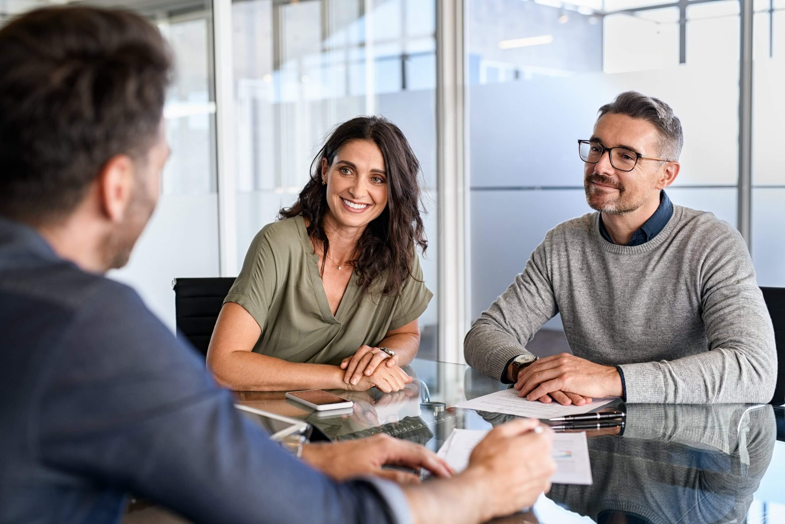 Three people sit at a glass table in a modern office, engaged in a friendly conversation about company culture. A woman and a man face another man whose back is to the camera. Papers, a smartphone, and eyeglasses are on the table. Three people sit at a glass table in a modern office, engaged in a friendly conversation about company culture. A woman and a man face another man whose back is to the camera. Papers, a smartphone, and eyeglasses are on the table.
