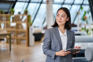 An employee in a gray suit stands in a modern office, holding a digital tablet and pen, looking to the side with a thoughtful expression. The background features stylish furniture and large windows.