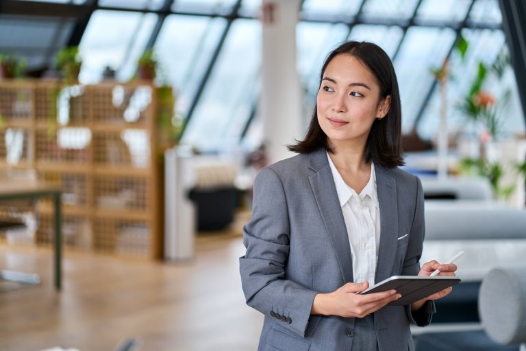An employee in a gray suit stands in a modern office, holding a digital tablet and pen, looking to the side with a thoughtful expression. The background features stylish furniture and large windows.