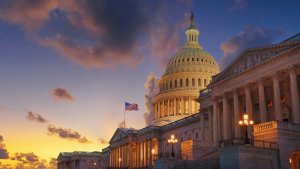 The U.S. Capitol building at sunset, with dramatic clouds in the sky, the dome of this iconic federal structure illuminated, and an American flag flying in front.