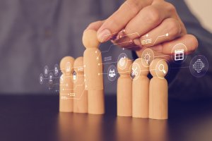 A hand arranges wooden human figures in a row, with digital business and technology icons overlaying the image, symbolizing concepts like teamwork, performance, management, and innovation.